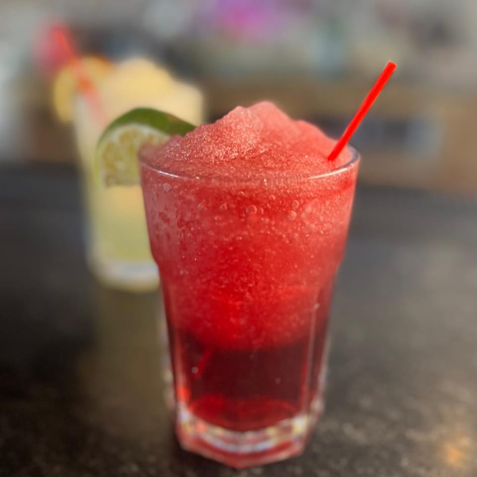 A vibrant red frozen drink with a red straw and a lime wedge, with a yellow drink in the background.