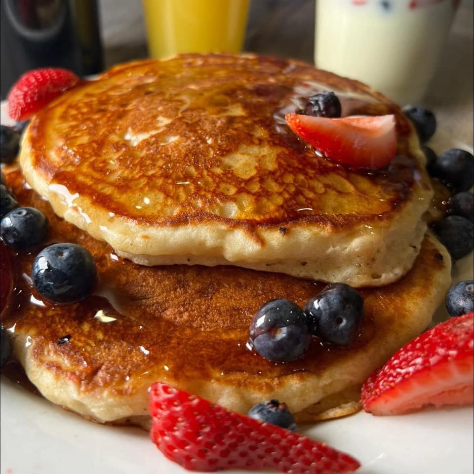 Stack of golden pancakes drizzled with syrup, topped with fresh blueberries and sliced strawberries.
