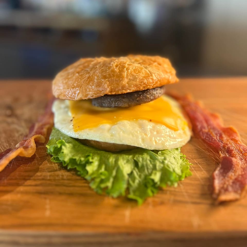 Breakfast sandwich with egg, cheese, sausage, and lettuce on a bun, with bacon strips.