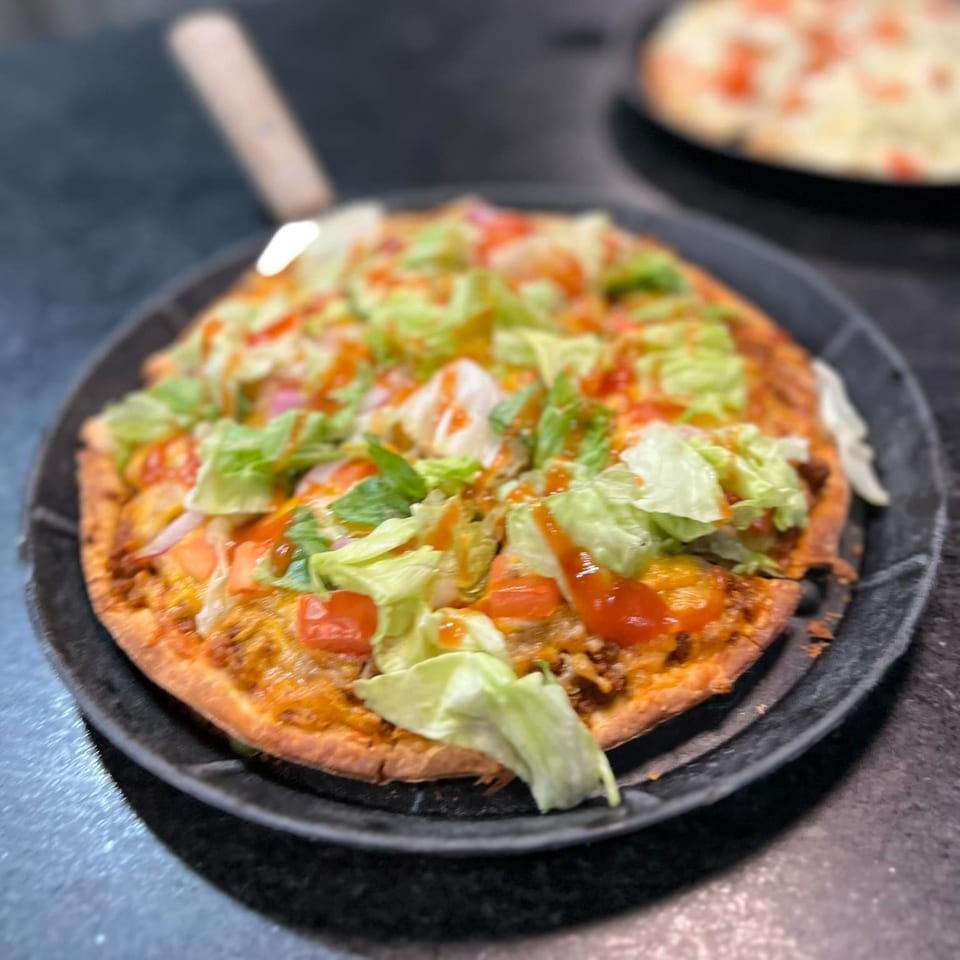 Roca Bar's taco pizza, topped with lettuce, tomatoes, and a drizzle of sauce, on a dark pan.